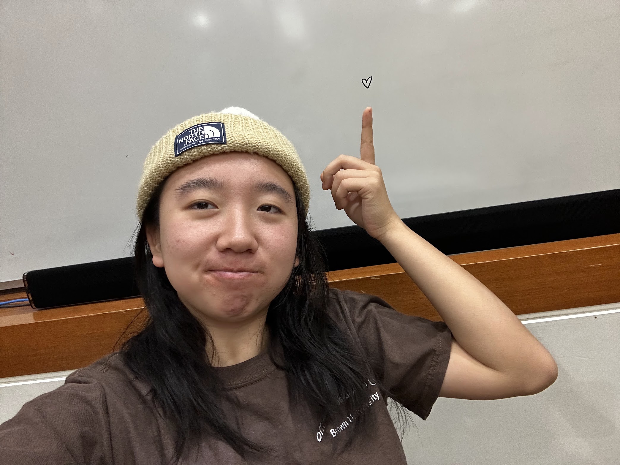 Selfie of Chilsea in a rice-colored beanie with a Brown University shirt. She is pointing to a whiteboard, where there is a heart drawn